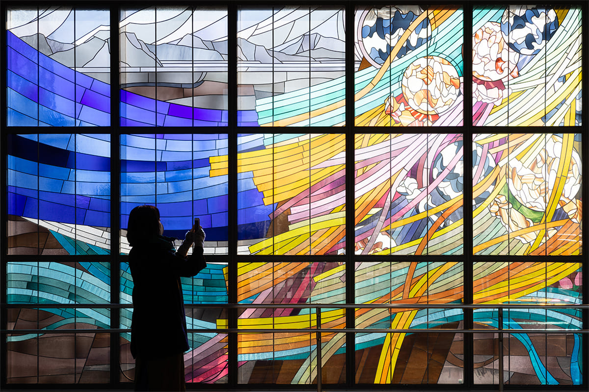 A person photographing the Stained Glass artwork “Beautiful water flow, dream in the blowing wind : FUSSA / AKISHIMA” at Haijima Station