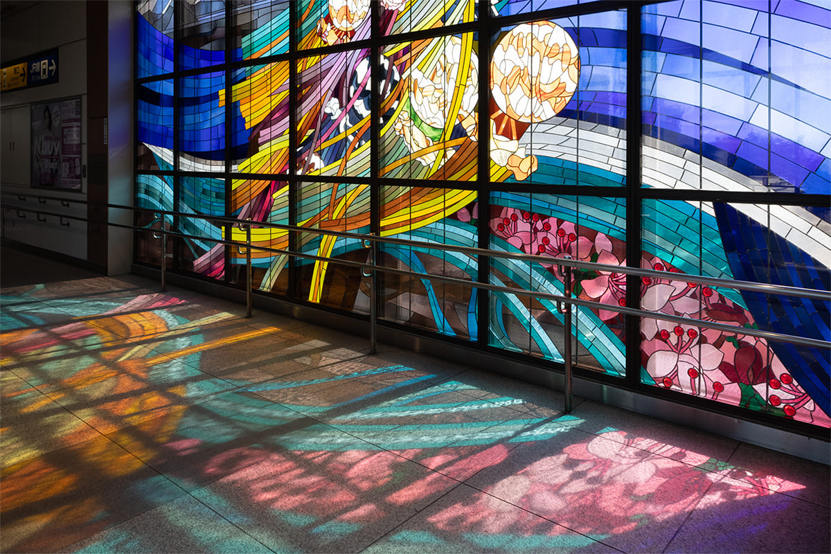The Stained Glass artwork “Beautiful water flow, dream in the blowing wind : FUSSA / AKISHIMA” illuminated by light at Haijima Station